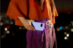 A person holding a smartphone with a light purple or lavender-colored back, partially placing it into their pocket. Photo Source: Google.com