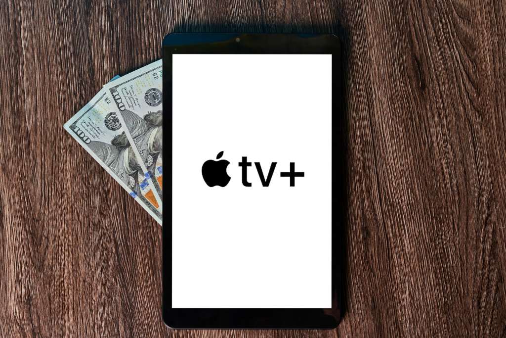 Apple TV+ logo displayed on a smartphone screen with U.S. dollar bills placed underneath on a wooden surface.
