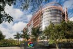 Modern curved glass Google office building in Bangalore with the Google logo sign visible at the entrance, surrounded by palm trees and tropical landscaping under a blue sky with white clouds.