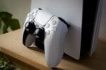 White PlayStation 5 DualSense controller resting against PS5 console on wooden shelf with plant in background