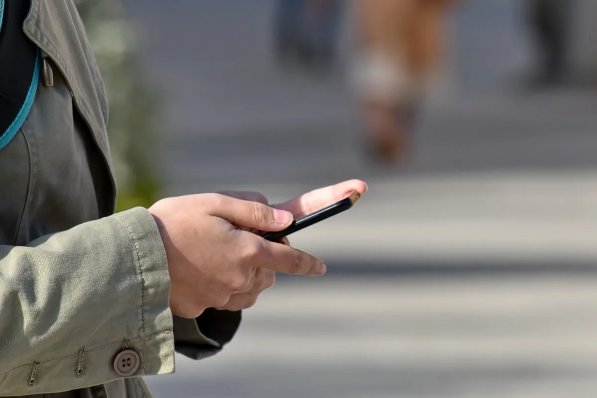 Close-up of person's hand holding a smartphone outdoors in an urban setting, wearing a gray jacket.