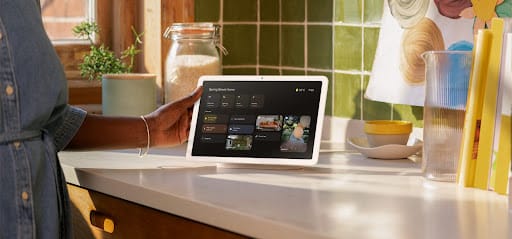 Google Nest Hub smart display on kitchen countertop showing the Google Home interface with photo gallery feature, surrounded by kitchen items including a glass vase and wooden canister.