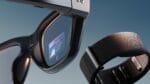 Close-up view of Meta Ray-Ban Display smart glasses showing the small color display in the right lens with a blue interface displaying Santorini travel information, positioned next to the black Neural Band wristband with textured surface.