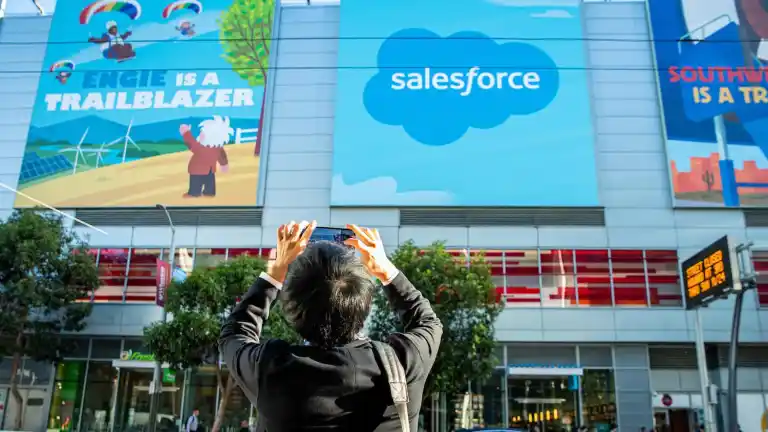 Person taking a photo of Salesforce headquarters building in San Francisco with the blue cloud logo prominently displayed on the facade alongside colorful murals stating "Eagle is a Trailblazer" and "South Beach is a Trailblazer"