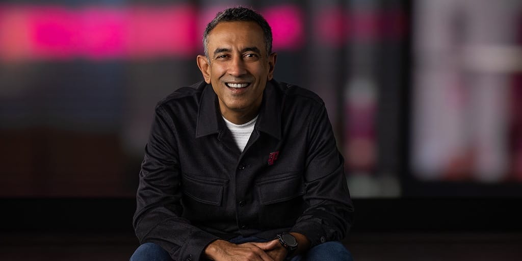 Srini Gopalan smiling in black T-Mobile branded jacket against blurred magenta background
