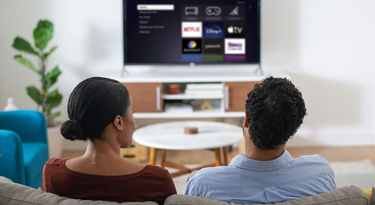 Two people on a sofa watching a TV with the Roku home screen of app tiles in a bright living room.